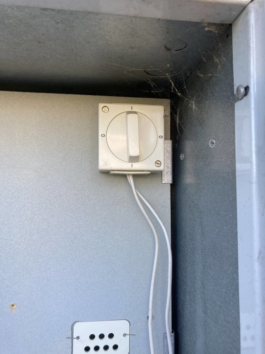 Elektrisk dosa monterad på en grå vägg med ett eluttag och sladd som leder nedåt, spindelnät syns i hörnet.