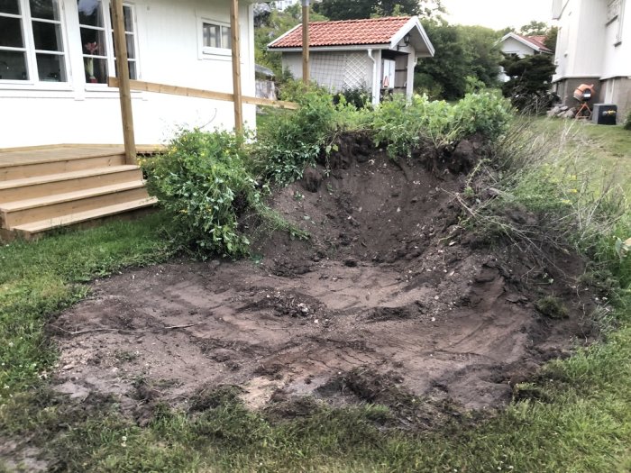 Uppgrävd backe vid vit husvägg med nybyggd trätrappa leder till dörr, intill blivande trappbygge.