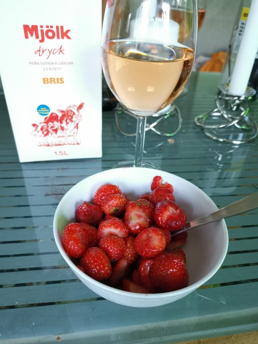 Skål med jordgubbar på bord bredvid ett glas rosévin och mjölkdryck-förpackning.