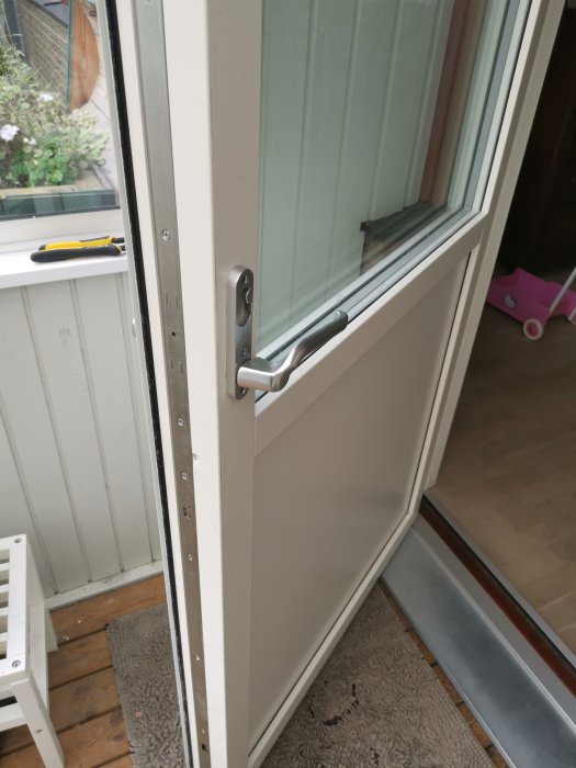 Halvöppen vit fönsterdörr med fokus på dörrhandtaget och synliga skruvhål i karmen, inuti ett hus.