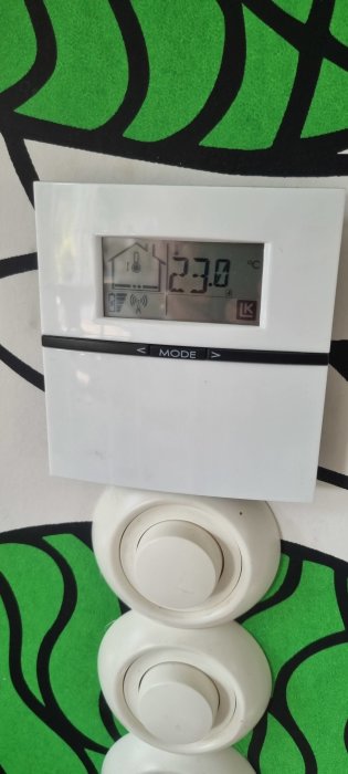 Termostat på vägg visar inomhustemperatur 23,8°C med färgfull bakgrund och del av ljusknapp under.