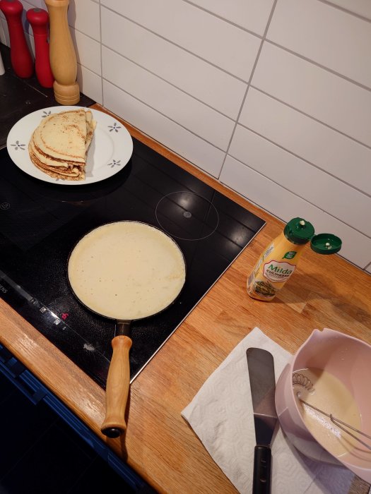 Tillagningsprocess av pannkakor med en stapel färdiga på en tallrik och smet i en bunke på spisen.