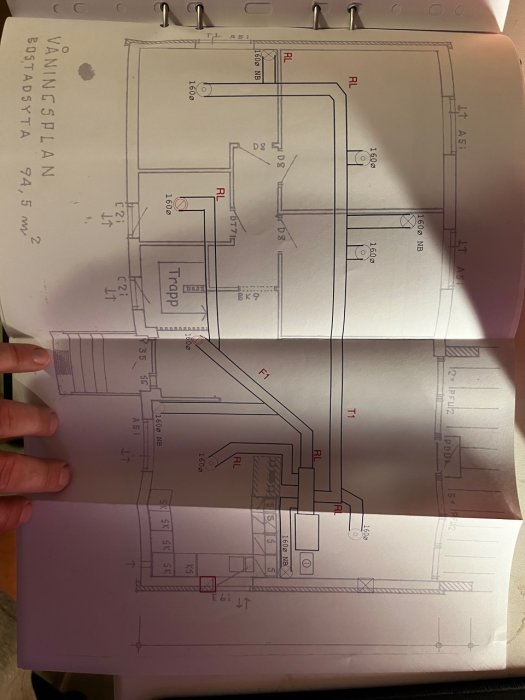 Hand håller upp öppen anteckningsbok med tekniska ritningar av VVS-installationer i en byggnad.