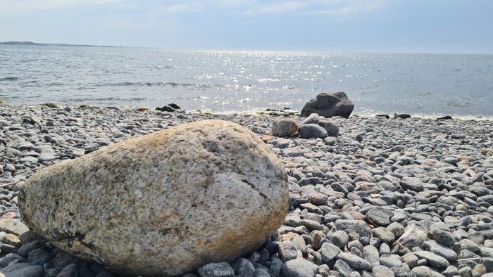 Stor sten i förgrunden på en strand med småsten och havet i bakgrunden, solkatter på vattnet.