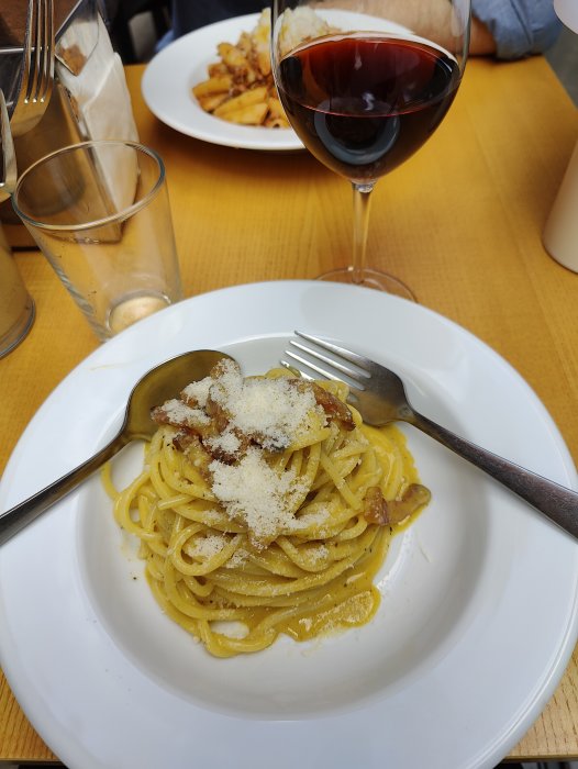 En tallrik pasta med parmesan och en sked, ett glas rött vin och en person som äter i bakgrunden.