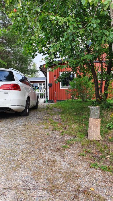 Vit kombibil parkerad på grusuppfart framför rött trähus med vita knutar och grönskande trädgård.