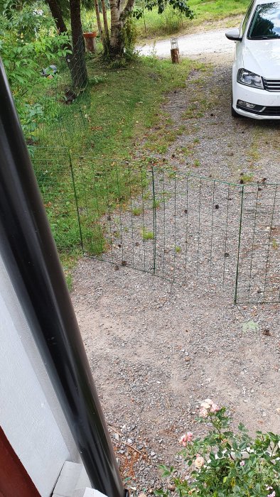 Utsikt över grusuppfart med grönt stängsel och parkerad bil sedd från ett fönster.