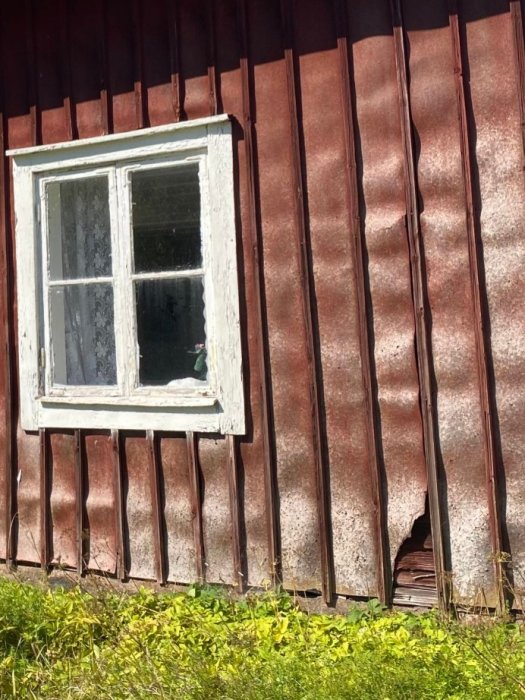 Del av en gammal byggnad med skadad röd plåtbeklädnad och synlig timmerstomme under ett fönster.