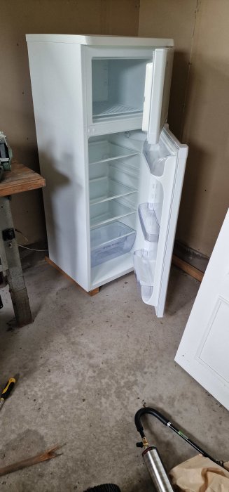 Tomt vitt kylskåp står öppet i ett garage med byggmaterial och verktyg omkring.
