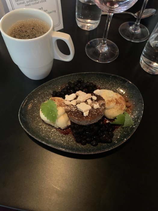 En tallrik med dessert bestående av två bakverk, grädde och bär, ackompanjerad av en kopp kaffe och ett tomt glas.