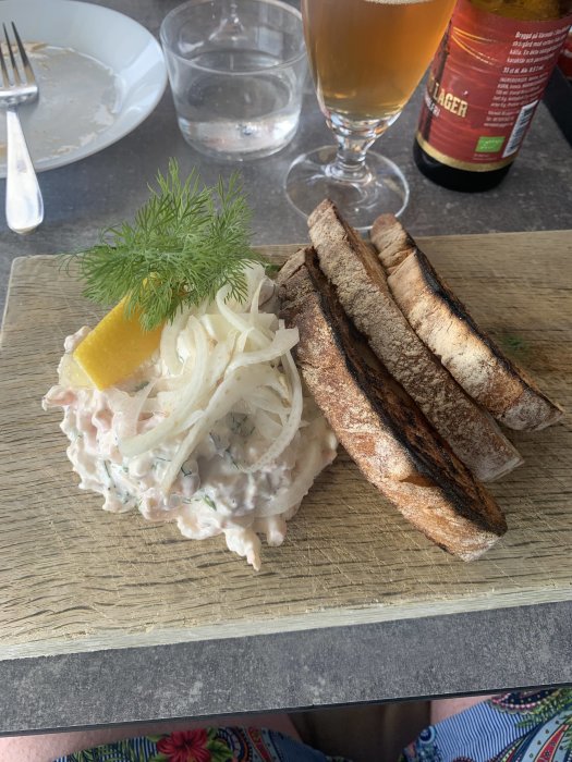 Skärgårdslunch på träplatta: Skagenröra, rostat bröd, öl och vatten, med dill och citrondekoration.