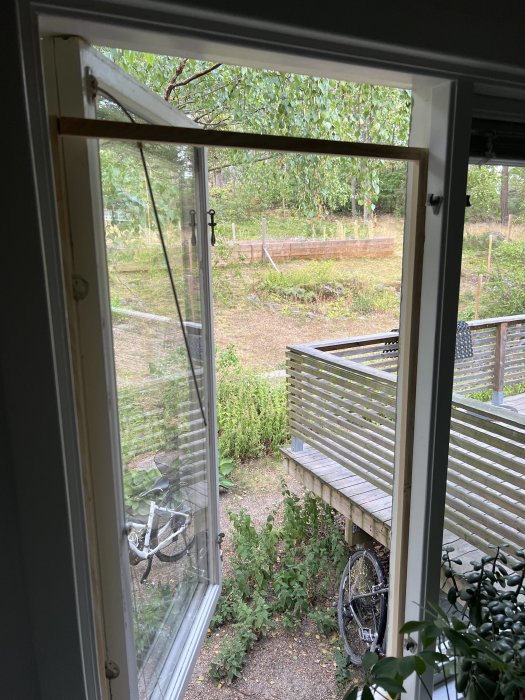 Öppet fönster med dåligt monterat grovmaskigt myggfönster, utsikt över trädgård och cykel.