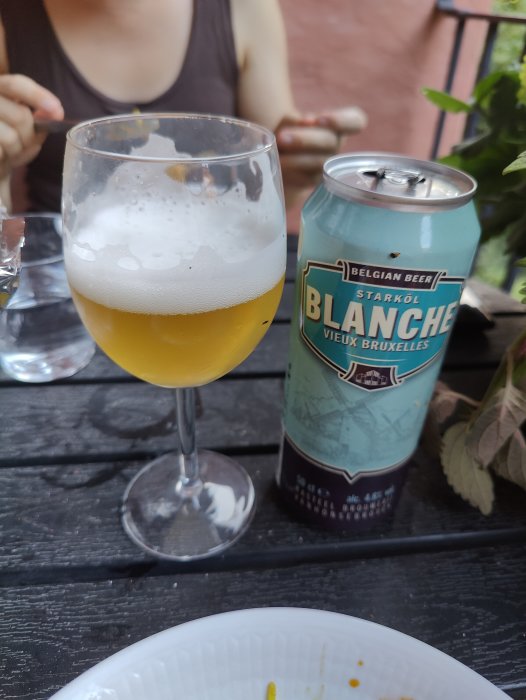 Ett glas veteöl och en ölburk på ett bord, med en suddig person i bakgrunden.