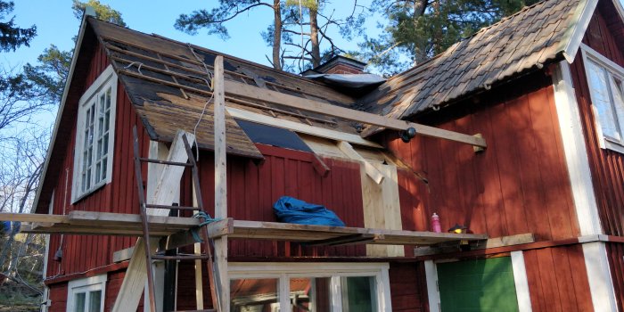 Renovering av rött torp med arbetsställningar, påbörjat takbyte och skog i bakgrunden.