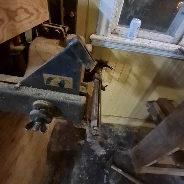 En snickarbänk i ett stökigt arbetsrum med spån och verktyg, nära ett dammigt fönster med en dryckesburk på fönsterbrädan.