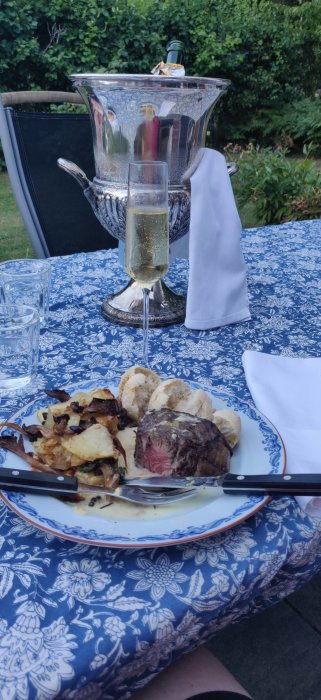 Stek med potatis och svamp, ett glas champagne och en vinkylare på ett dukat bord utomhus.