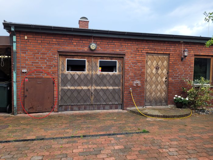 Garage med tegelfasad och trägarageport, husdörr och gammal elkabelskåp markerad med röd cirkel.