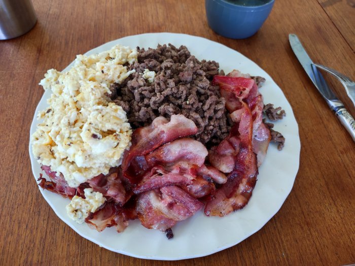 Tallrik med äggröra, stekt bacon och köttfärs serverad på ett träbord med bestick.