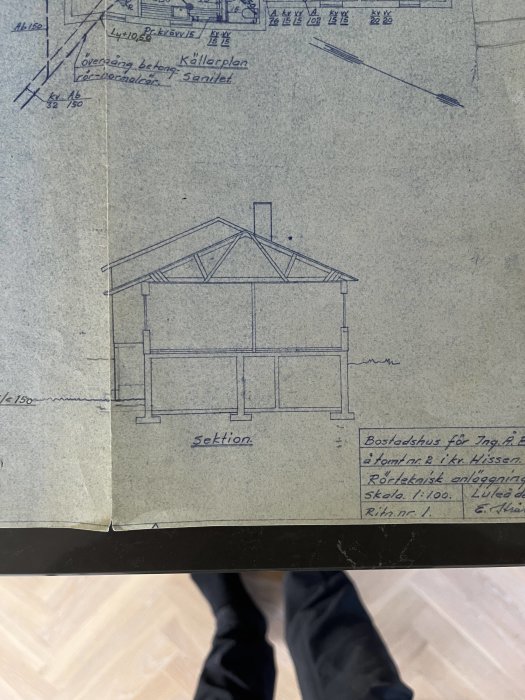 Ritning av en byggnadssektion som visar ett bostadshus med måttagningar och noteringar, placerad på ett golv med synliga fötter i nederkanten.