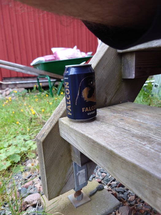 En burk Falcon Export öl står på en trätrall nära justerbar stolpsko med en skottkärra i bakgrunden.