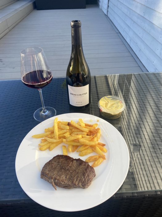 En tallrik med biff och pommes frites, ett glas rödvin och en flaska vin, bredvid bearnaisesås, på ett utebord.