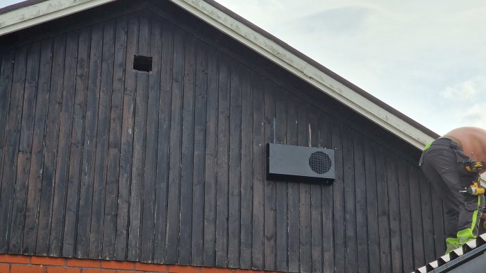 Arbetare på stege installerar ventilationssystem på mörkt trähusvägg med öppen lucka ovanför.