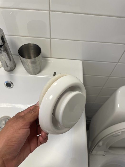 Hand håller en demonterad toalettsits framför en toalettstol i ett kaklat badrum.