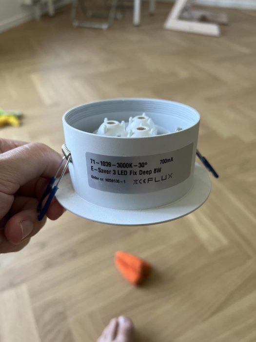 En hand håller en LED-downlightlampa mot ett golv med trätextur i bakgrunden.