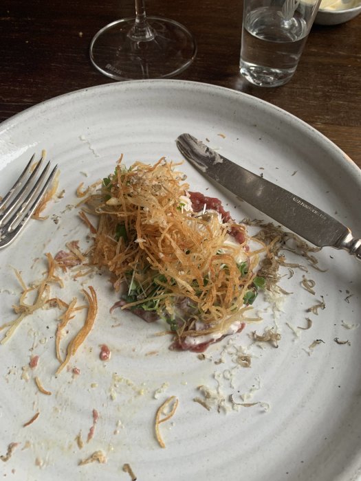 Halväten portion av oxtartar med krispig potatis och tryffelkräm på en vit tallrik med bestick.