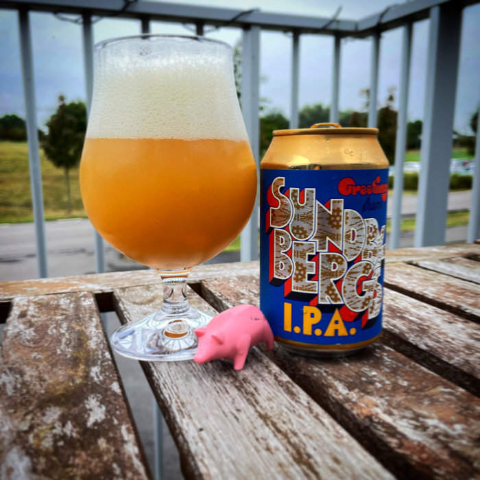 Ölglas fyllt med skummande öl på träbord vid balkong med utsikt, färgglad ölburk och liten rosa gris leksak.