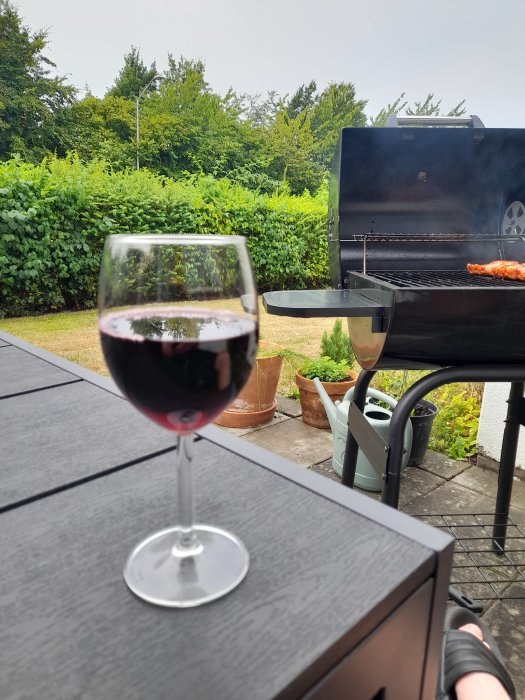 Ett glas rödvin i förgrunden med en grill i bakgrunden på en uteplats.