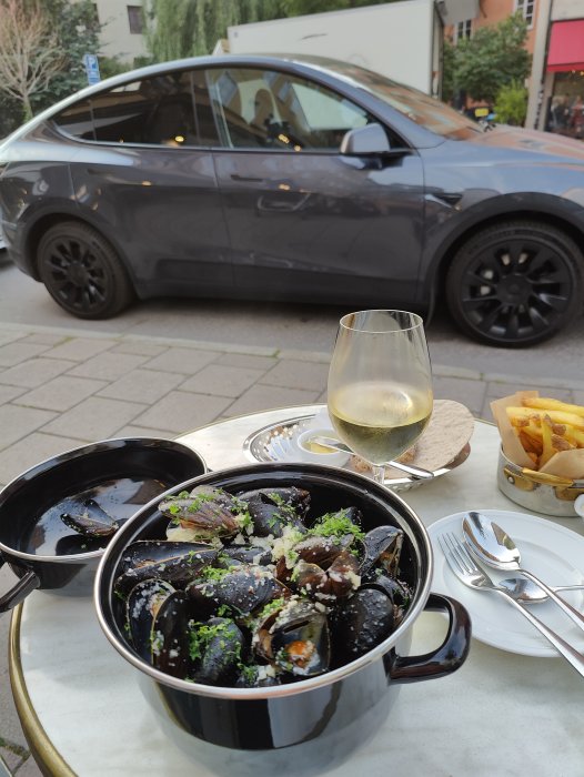 En gryta med musslor, ett glas vin och pommes frites på en restaurangterass med en bil i bakgrunden.