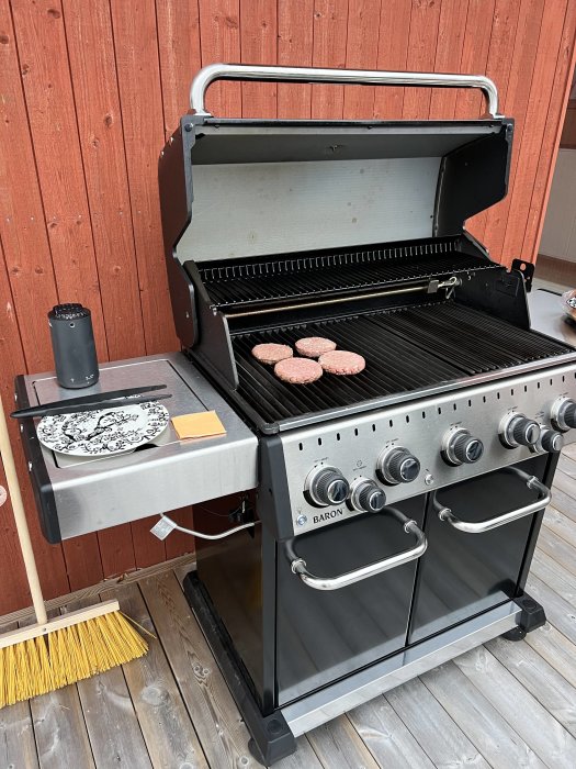 Gasolgrill på träaltan med hamburgare på grillgallret, en tallrik vid sidan och en sopborste i bakgrunden.