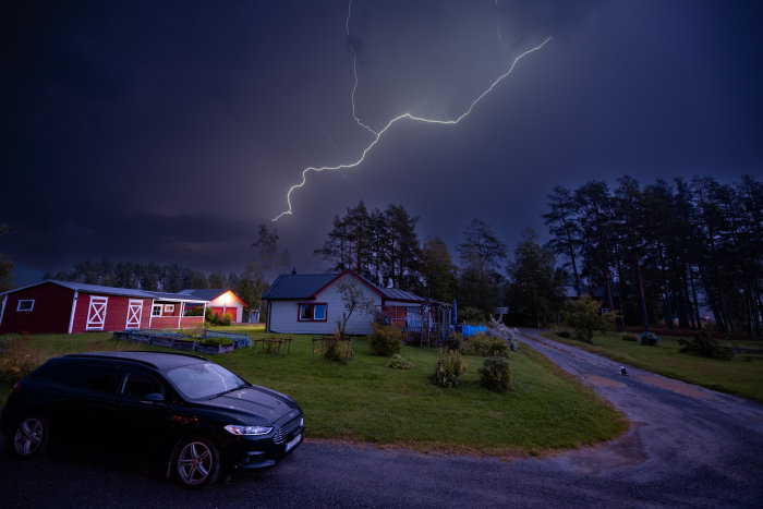 En blixt lyser upp himlen över ett landskap med hus och bil, katt synlig i förgrunden.