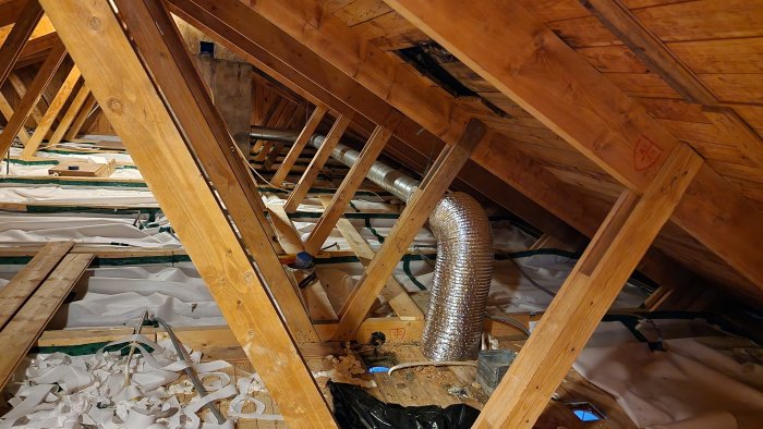 Flexibel metallkanal för ventilation som sträcker sig genom en vind med träbjälkar och isoleringsmaterial.