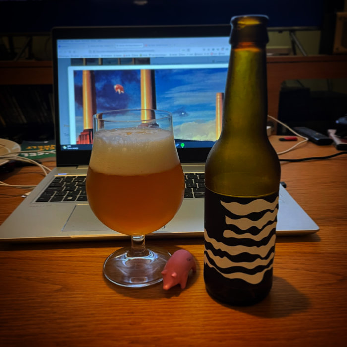 Ölglas med skum vid en laptop, ölflaska med randig etikett och en liten rosa figur intill.