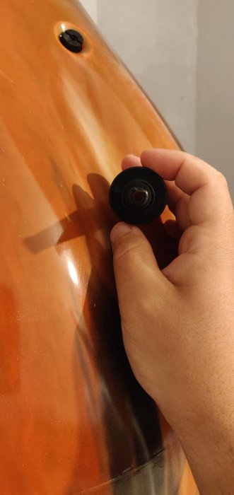 Hand håller ett svart föremål mot en orange yta för att visa dess storlek, ungefärligen 1.2 cm i diameter.