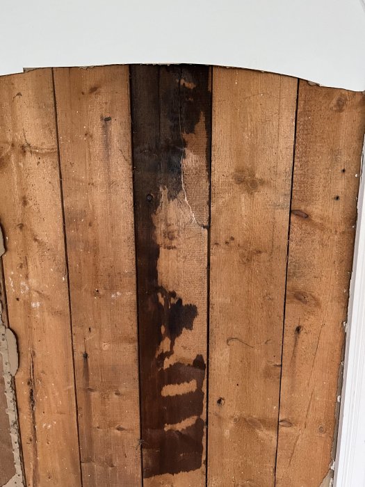 Bruna trägolvplankor med vattenfläckar och skador vid en vit väggkant.