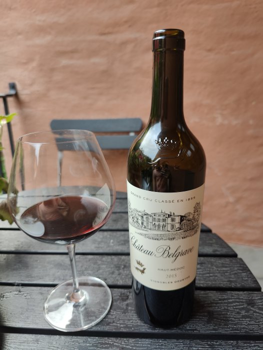 Flaska Château Belgrave 2015 Bordeaux vin bredvid ett halvfullt vinglas på ett träbord.