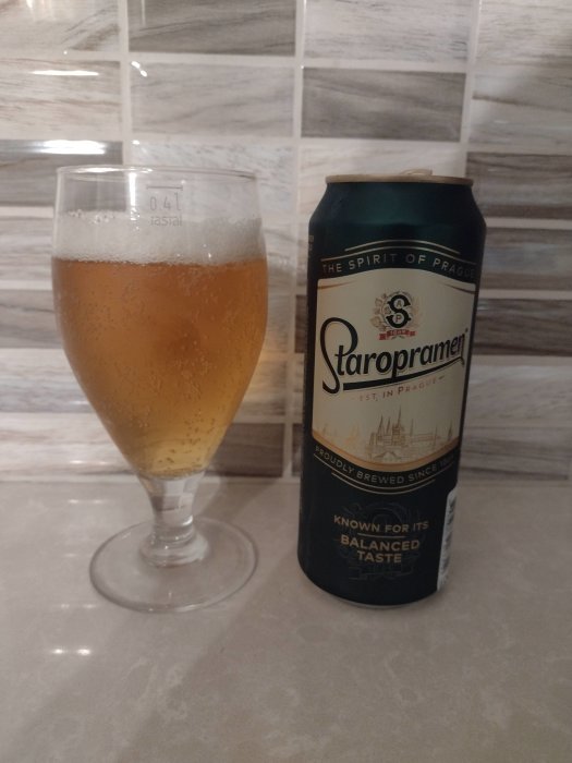 En Staropramen ölburk bredvid ett glas fyllt med öl på en bänkskiva framför kakelvägg.