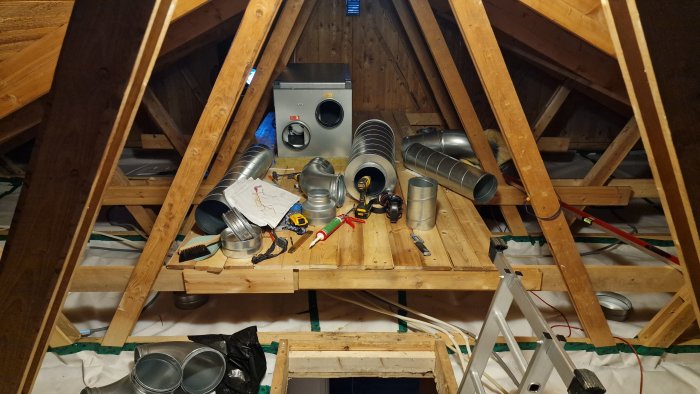 Ventilationskanaler och verktyg på ett trägolv i en vind med öppna takstolar.