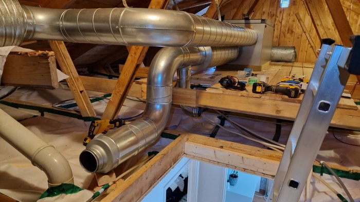 Ventilationsrör på vinden med isolering och verktyg synligt på träbjälkar samt en öppen dörr nedtill.