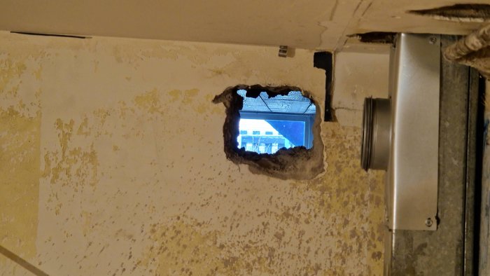 Hål i en vägg med synligt ventilationsrör och utsikt till en arbetsplats utanför.