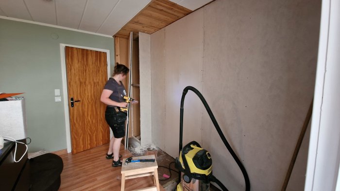 Person monterar gipsvägg med skruvdragare bredvid en dammsugare och arbetsmaterial.