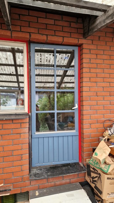 Begagnad blå dörr med fönster insatt i röd tegelvägg, med ommålning och uppsättning av lister pågående.