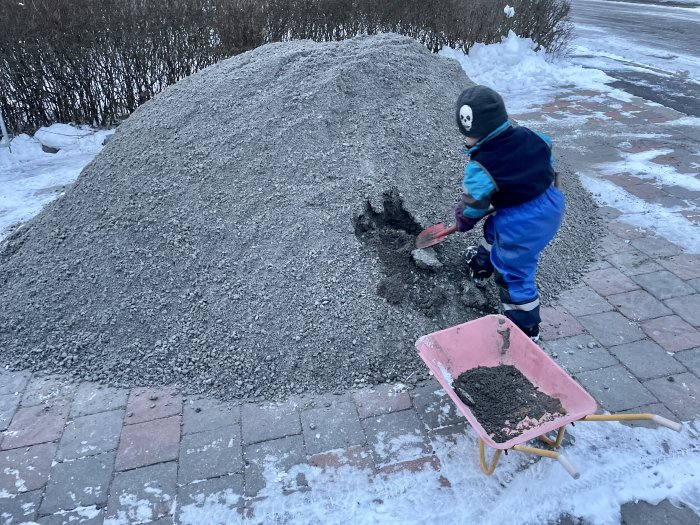 Barn i vinterkläder hjälper till med trädgårdsarbete genom att skotta grus i en skottkärra.