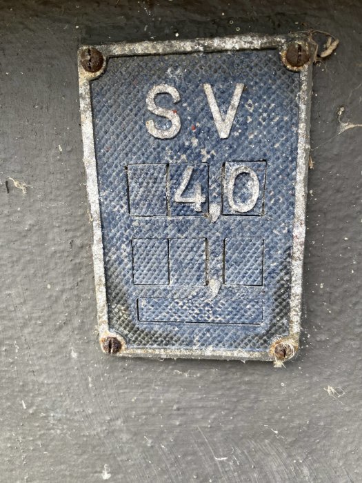 Gammal blå skylt med texten "SV 410" monterad på en grå vägg.