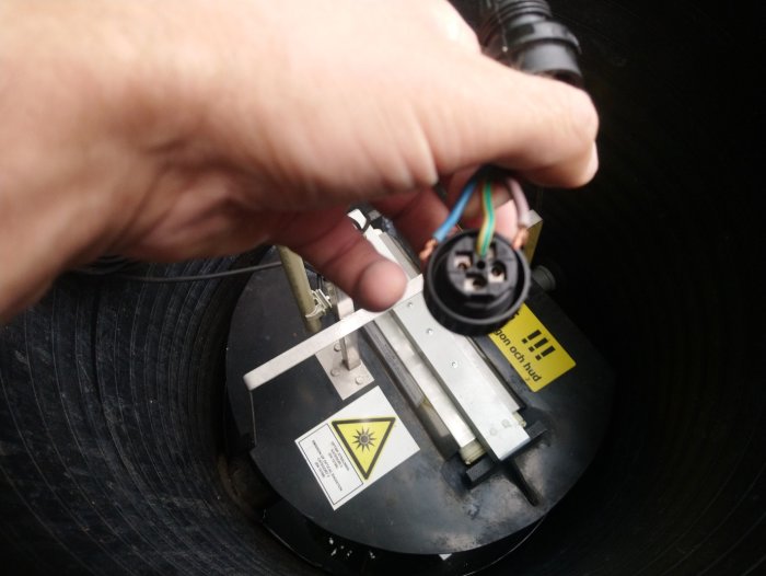 Hand håller en elektrisk kontakt med kablar framför en öppen anordning med varningsetiketter.