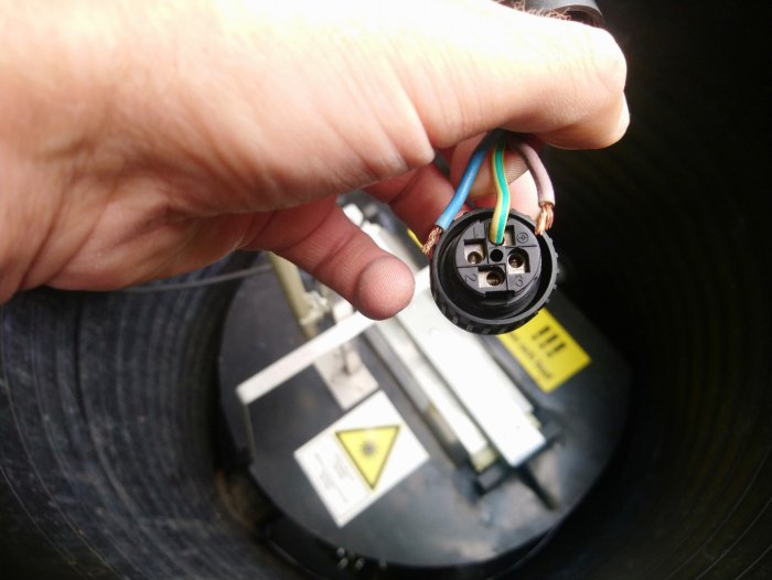 Hand håller ett elektriskt kopplingsuttag med synliga trådar framför en oskarp bakgrund med varningsetiketter.