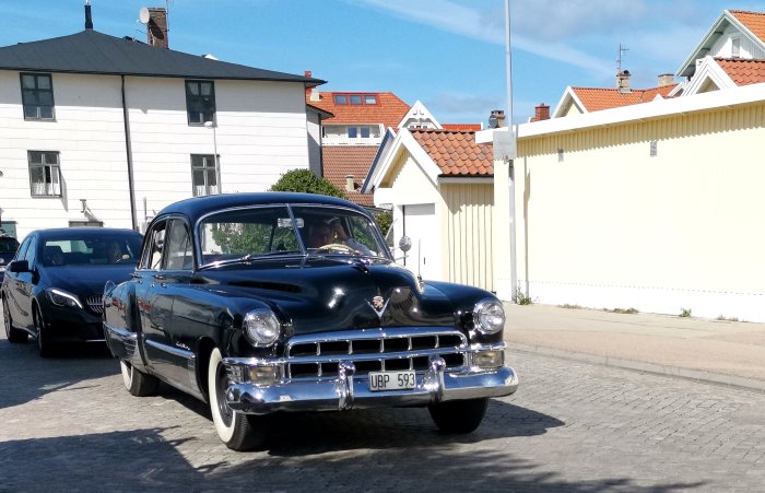 Klassisk svart bil från 1950-talet kör på gata med hus i bakgrunden.